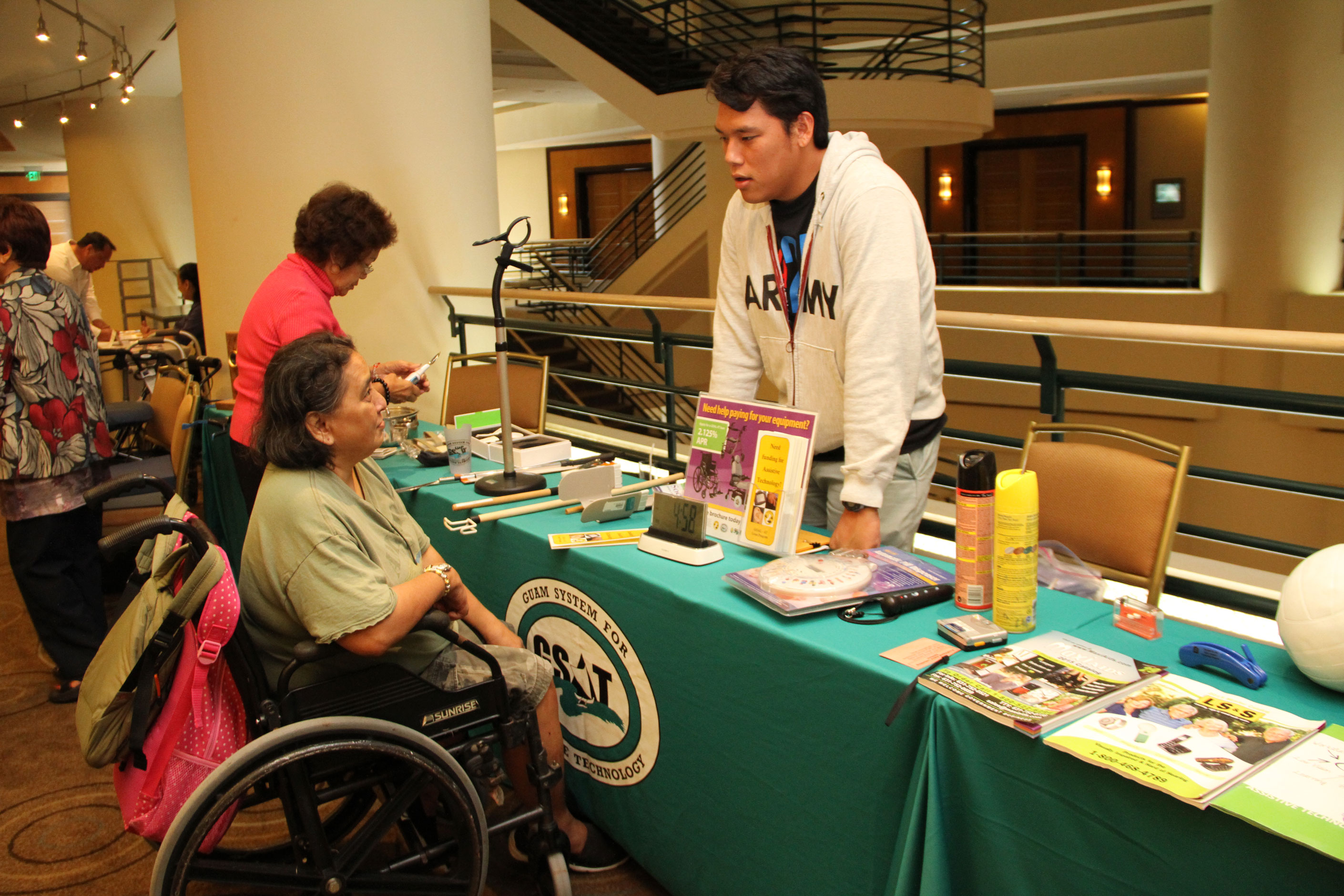Individual in wheelchair speaking with individual standing behind table with various assistive technology devices.